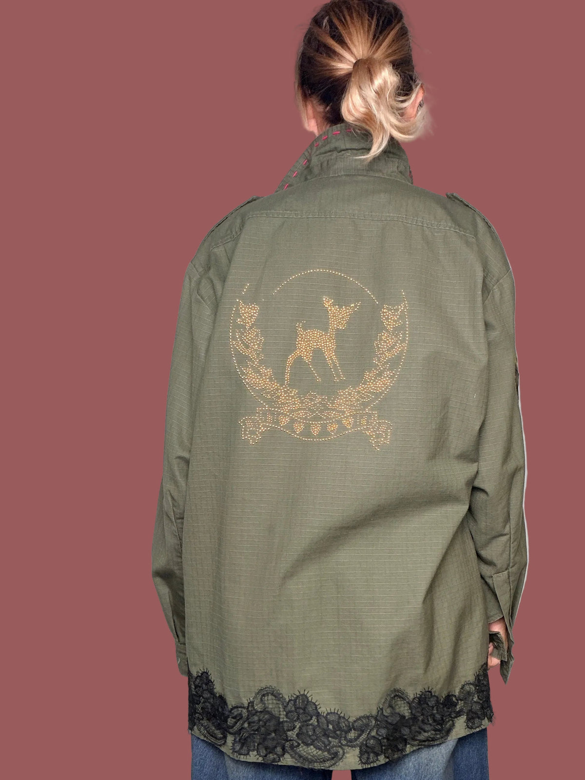 Giacca militare verde oliva con patch decorativi e pizzo nero sul fondo, vista frontale con braccia aperte